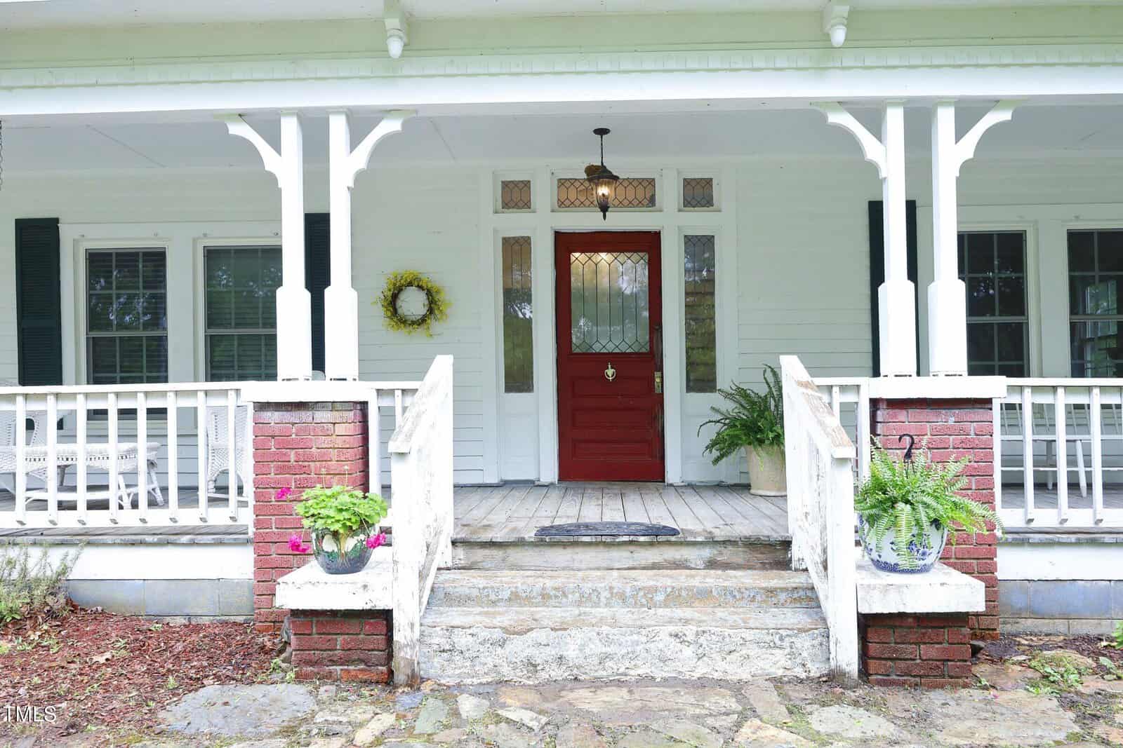 A white house with a covered front porch, red brick columns, and a red front door. There are potted plants on either side of the steps and a yellow wreath on the left window. The porch has white railings and trim.
