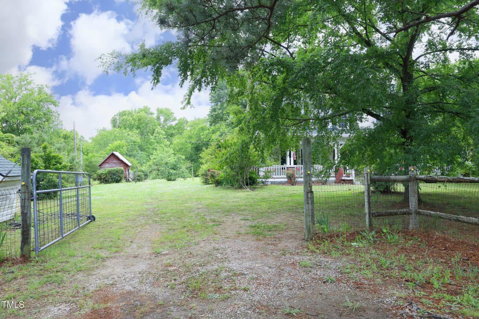 A gated entrance opens to a grassy yard with scattered trees, a wire fence, and a small building in the distance. Part of a house and front porch are visible on the right side. The sky is partly cloudy.