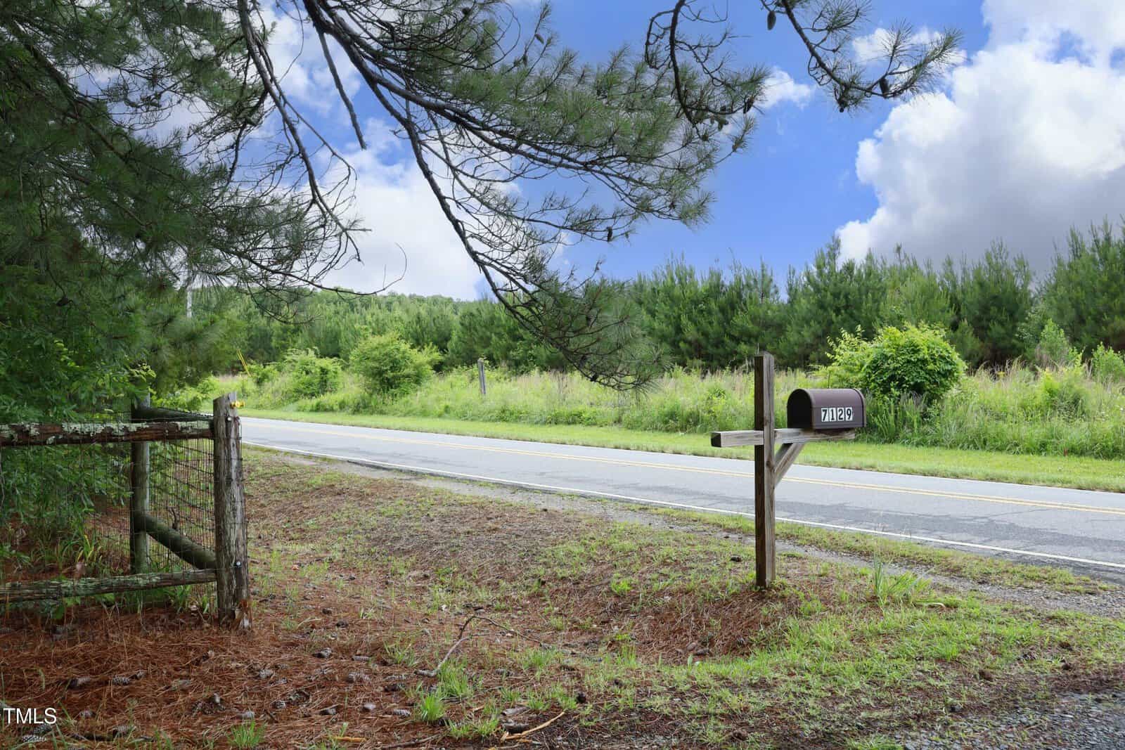 A rural roadside scene with a black mailbox on a wooden post, a metal gate with fencing, and a paved road bordered by grass and trees under a partly cloudy sky.