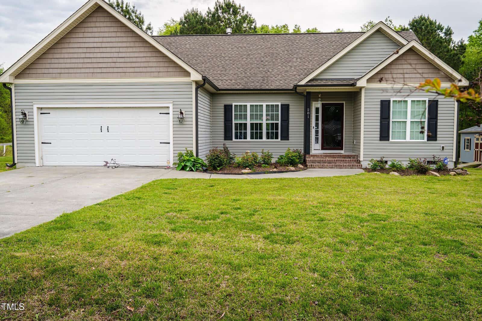 Charming single-story suburban house at 201 Lexington Park Drive in Oxford, featuring gray siding, a gable roof, attached two-car garage, white trim, black shutters, small front porch, and a well-kept lawn with landscaped bushes and plants.