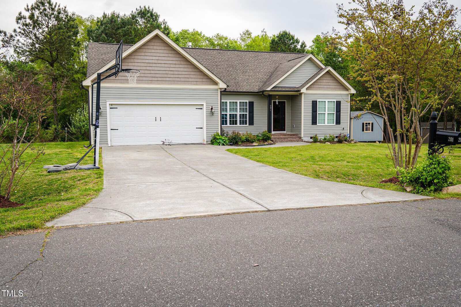 Single-story house with gray siding, a two-car garage, a basketball hoop, and a wide driveway. The front yard has grass, shrubs, young trees, and a small shed is visible in the backyard.