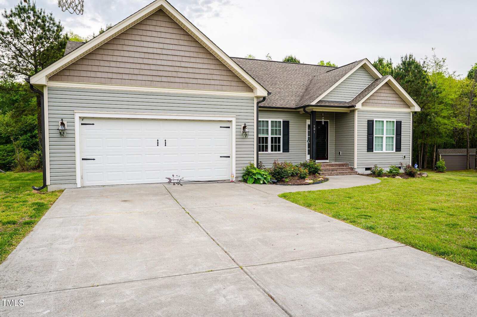 A single-story house with gray siding, a double garage, front steps leading to the entrance, and a concrete driveway. The yard is landscaped with grass, shrubs, and small trees.