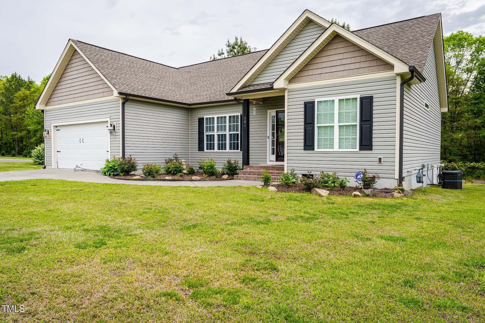 Single-story house with light gray siding, dark shutters, two-car garage, and a covered front porch. The yard has green grass and small shrubs planted along the front of the house. Trees are visible in the background.