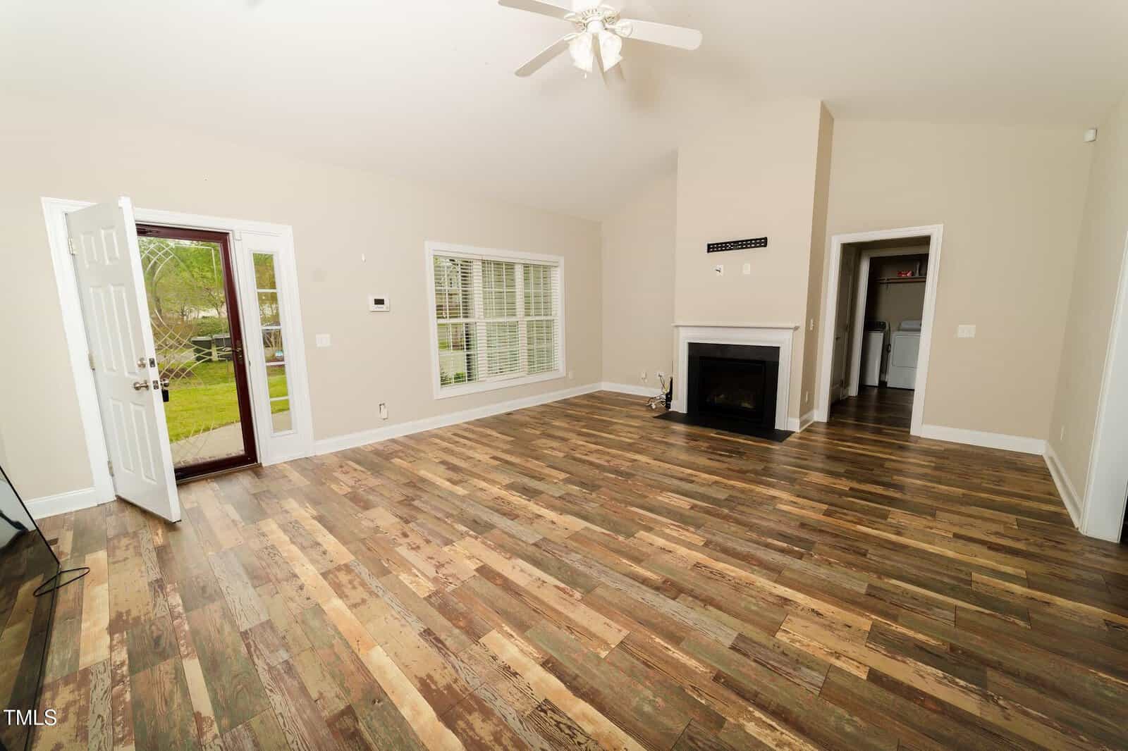 Spacious empty living room with wood-patterned flooring, light beige walls, a ceiling fan, fireplace, and large windows. The front door is open, revealing a view of grass outside. A kitchen is visible through a doorway.