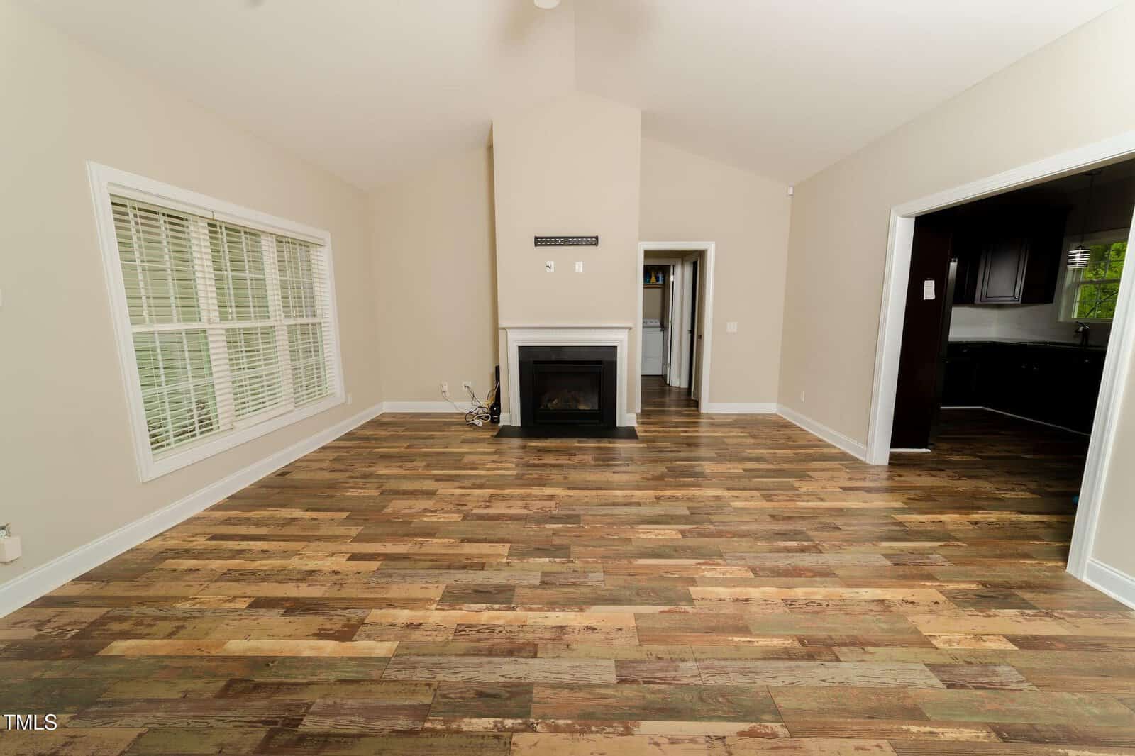 Spacious empty living room with patterned wood flooring, beige walls, large white-framed windows on the left, and a central fireplace; two doorways lead to adjacent rooms.