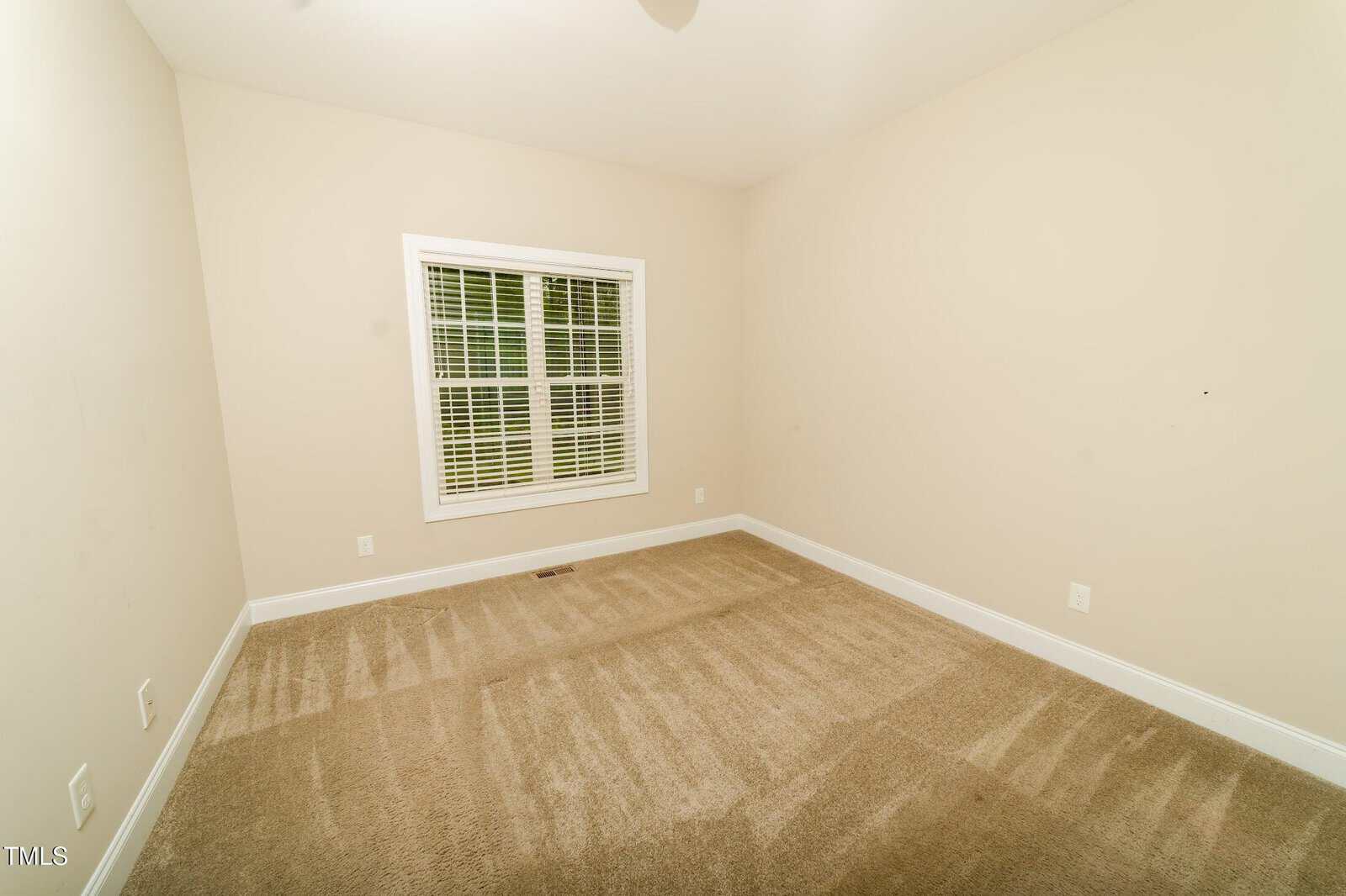 A small, empty room with beige carpet, white baseboards, off-white walls, and a window with white blinds letting in natural light. The floor shows vacuum lines and there are multiple electrical outlets.