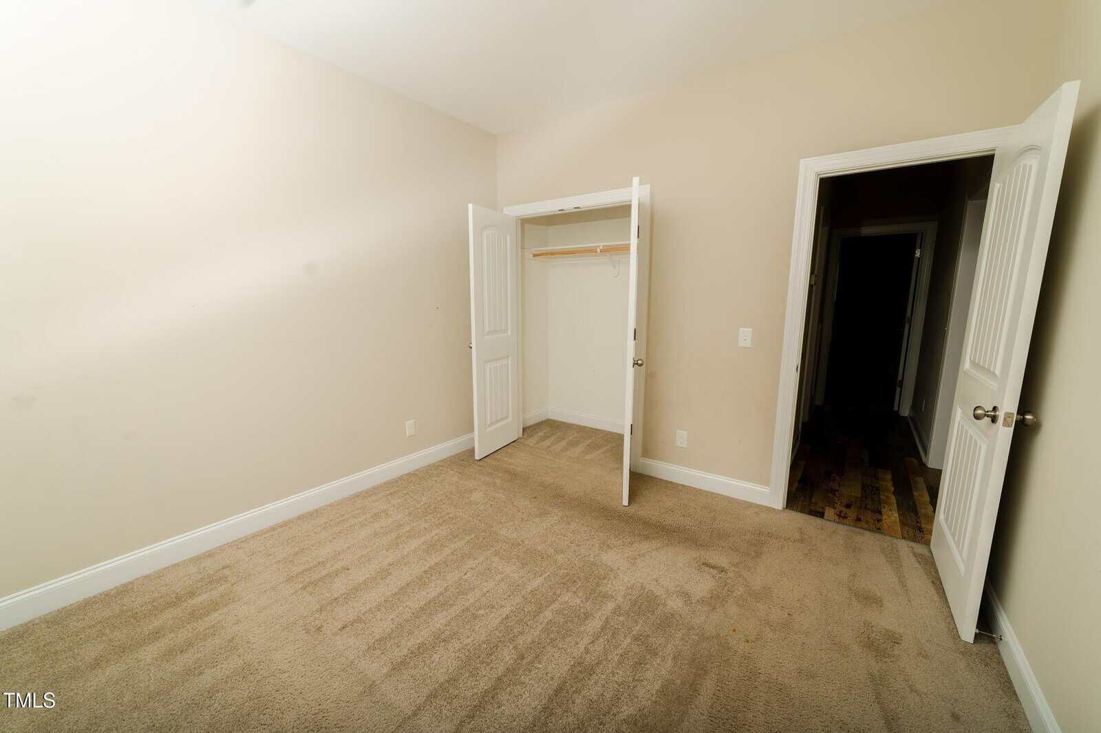 Empty room with beige carpet, cream-colored walls, an open closet with white double doors, and an open door leading to a hallway with wood flooring. The room is well-lit and has no furniture.