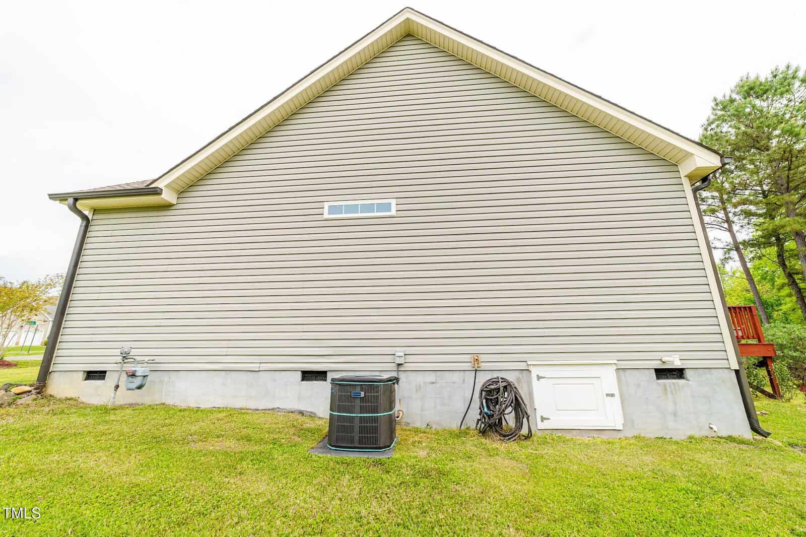 The exterior side of a house with beige vinyl siding, a small horizontal window near the roof, an air conditioning unit, a hose reel, utility meters, and a white access door on a concrete foundation. Grass covers the ground.