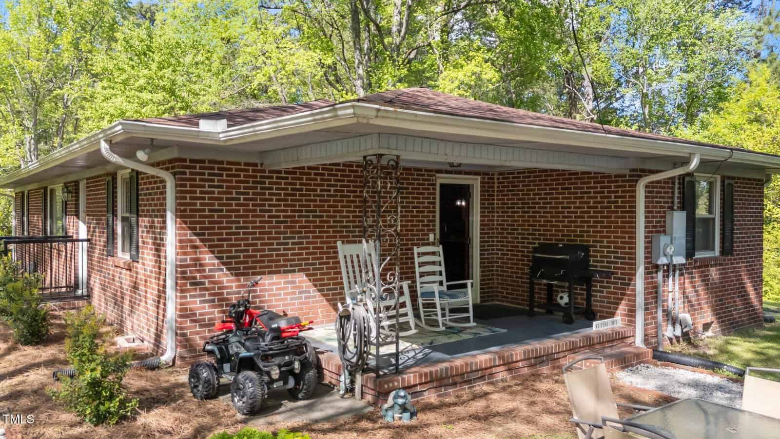 A small brick house with a covered back porch, two white rocking chairs, a grill, and a child's toy vehicle on the grass. The yard is surrounded by trees and features outdoor seating.