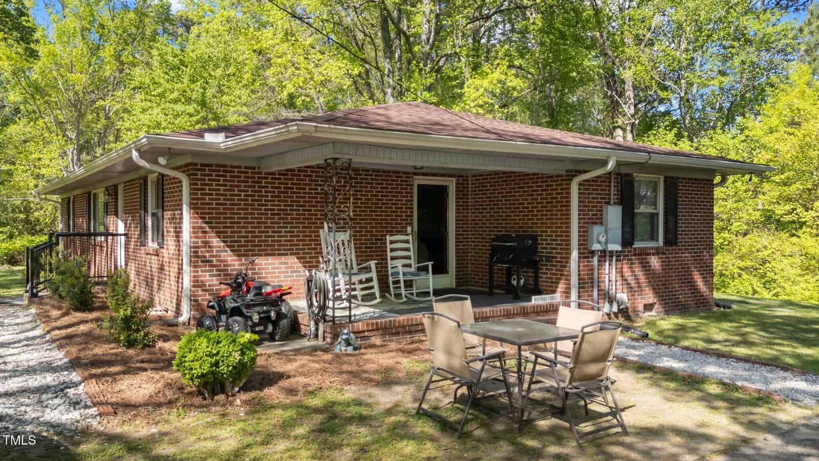 A one-story brick house with a covered porch featuring white rocking chairs and a grill. A glass patio table with four chairs is on the lawn. Trees and shrubs surround the house, and a small toy vehicle is near the porch.