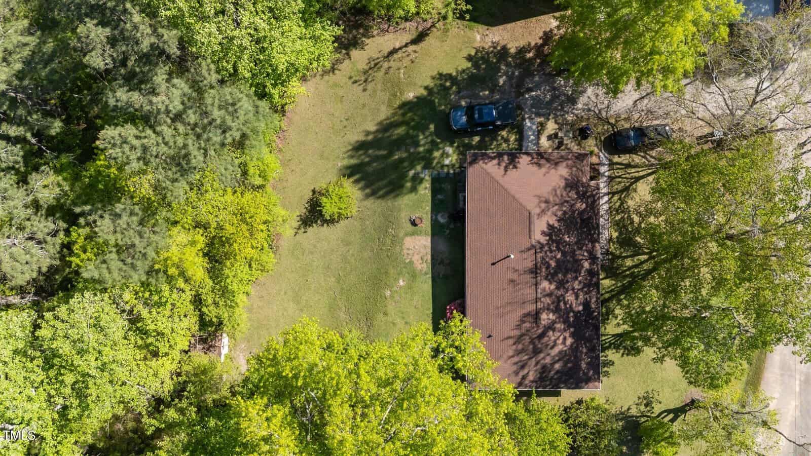 Aerial view of a house with a brown roof surrounded by green trees and grass. Two cars are parked on a driveway to the right of the house. Shadows from trees are visible on the lawn and roof.