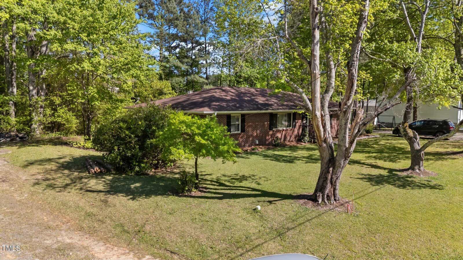 Single-story brick house with a brown roof set among mature trees, surrounded by a grassy yard. The home sits back from the street with some shade provided by trees in the front yard.