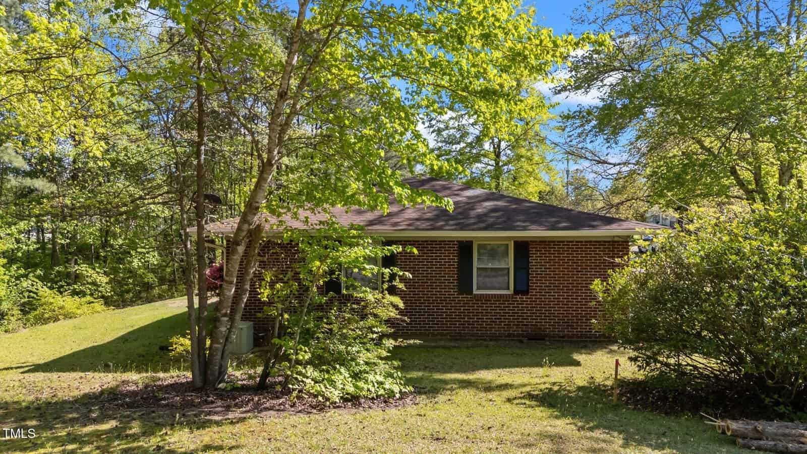 A one-story brick house with black shutters is partially obscured by trees and shrubs in a grassy yard on a sunny day. The roof is dark and the sky is clear with minimal clouds.