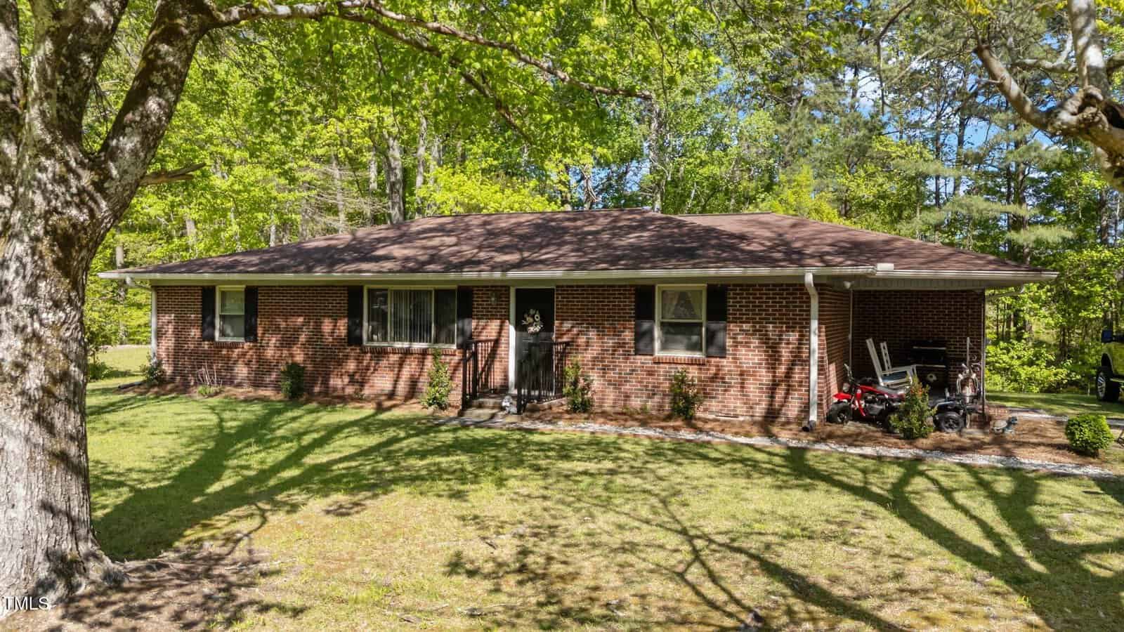 A single-story brick house at 906 Circle Drive, Creedmoor, features a brown shingle roof and sits on a grassy lawn shaded by large trees. Small shrubs accent the front, with items like a bicycle near the right side of the home.