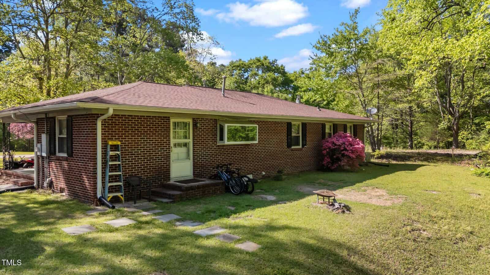 A single-story brick house with a brown roof sits in a grassy yard bordered by trees. There is a small set of steps, a white back door, a bicycle, a ladder, and a bush with pink flowers near the house.
