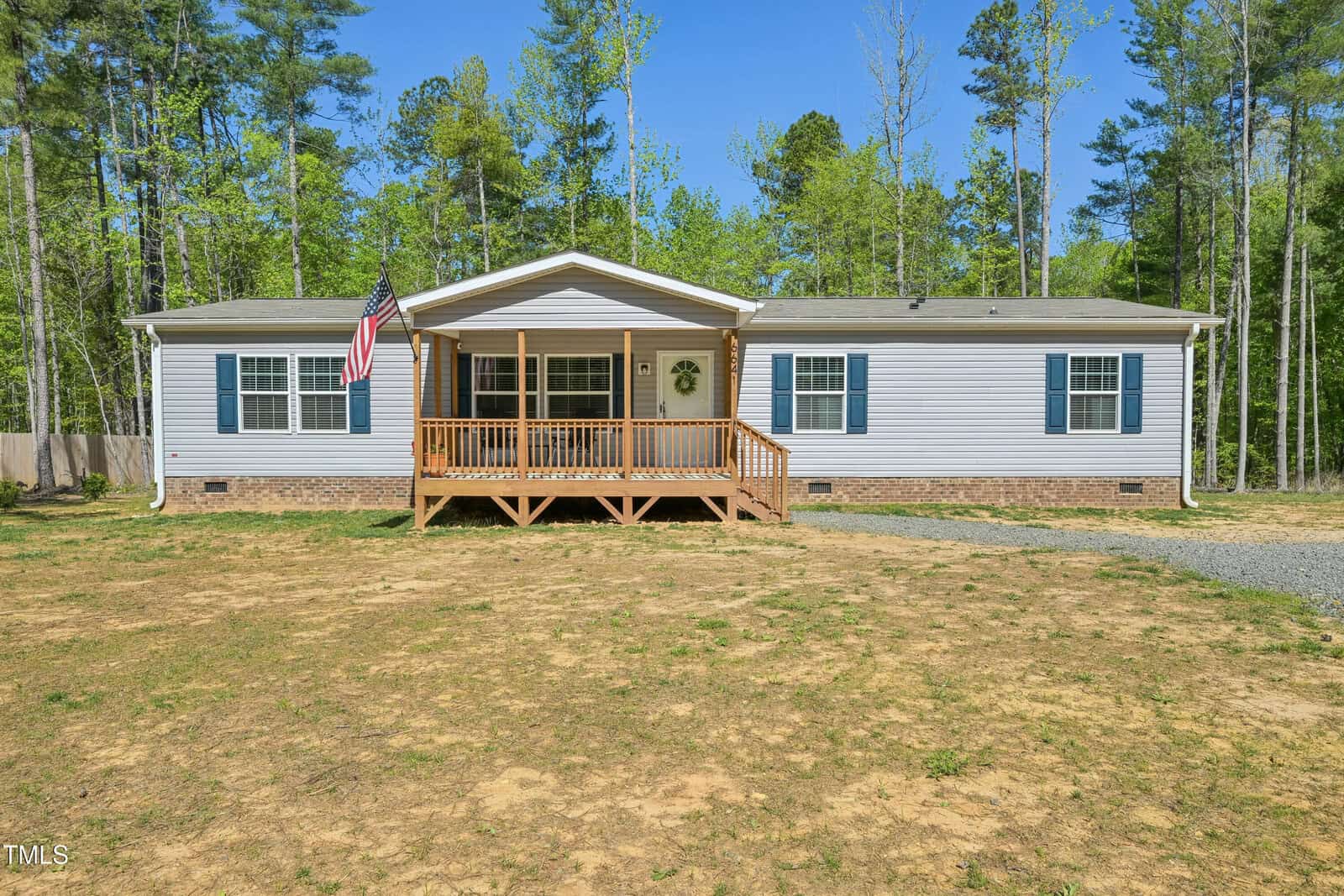 A single-story manufactured home at 6641 Woodlawn Drive, Oxford, with gray siding, blue shutters, and a small front porch with wooden steps. An American flag hangs by the entrance. The house sits on a patchy lawn surrounded by trees.