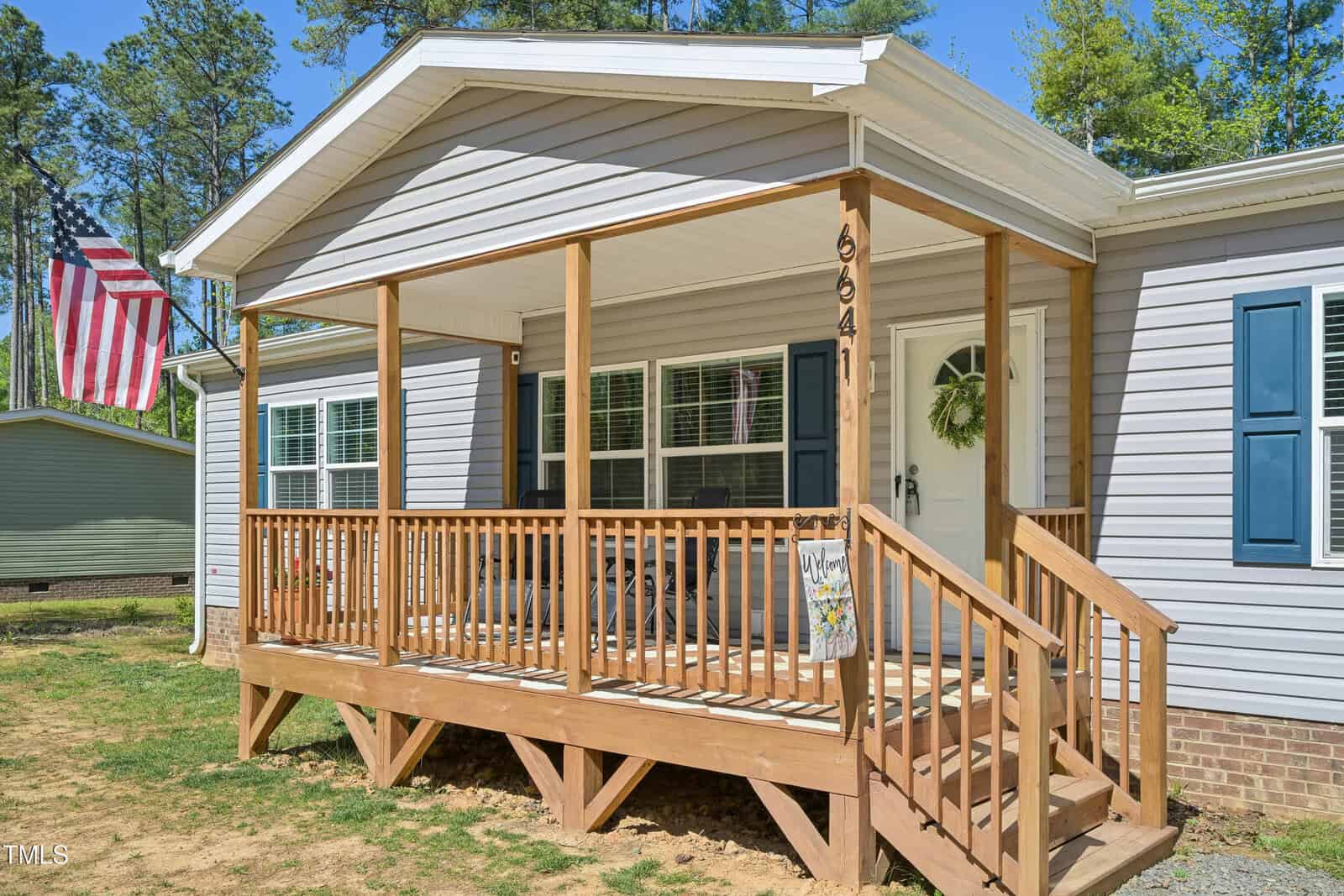 A light gray single-story house with a covered wooden front porch, white trim, dark blue shutters, and an American flag on a pole in the yard. The porch has a railing and a small welcome sign by the door.
