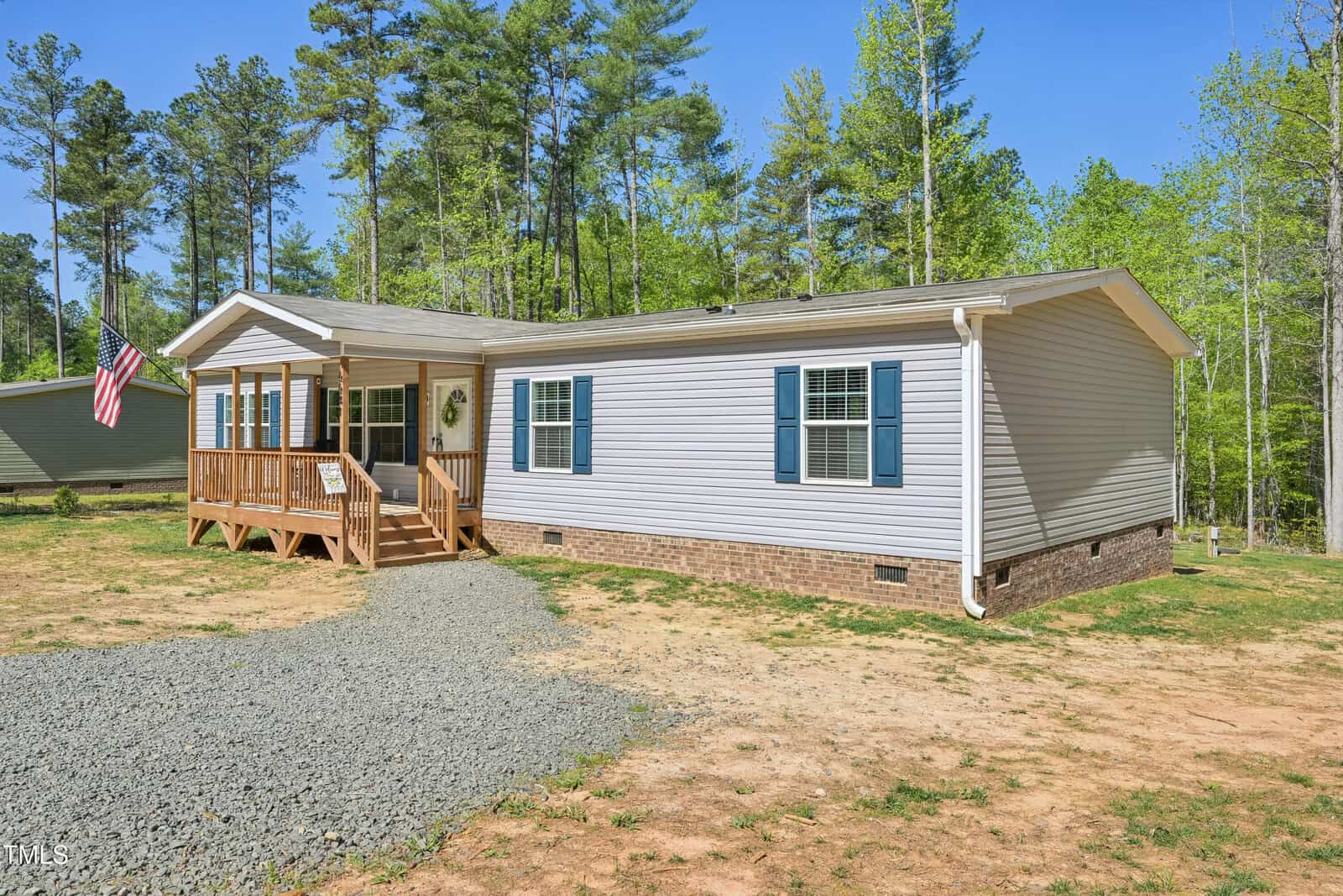 Single-story manufactured home with light gray siding, blue shutters, a small front porch with a wooden railing, set on a brick foundation, surrounded by trees and a gravel driveway. An American flag is on the porch.