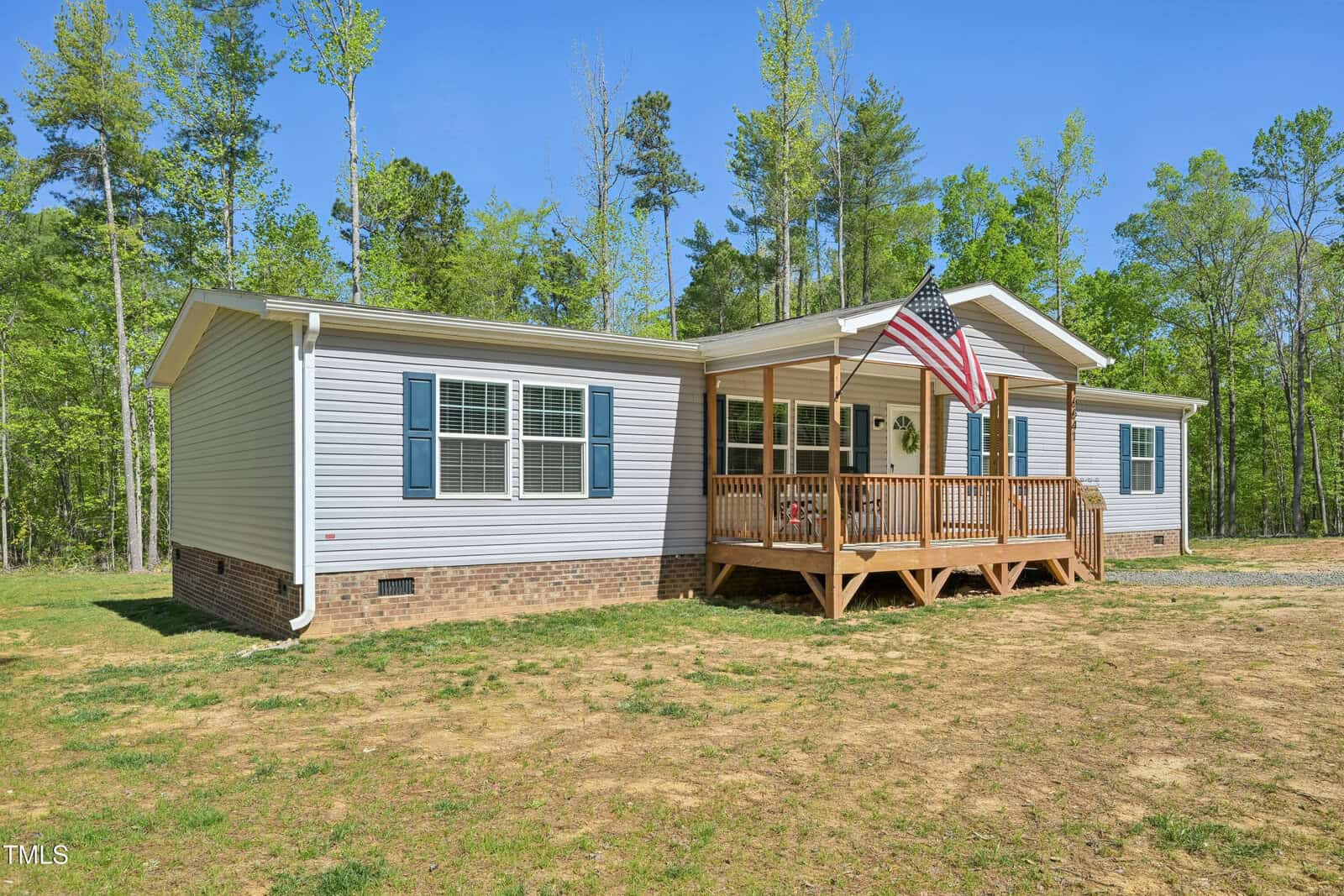 Single-story house with light-colored siding, brick foundation, blue shutters, and a covered front porch. An American flag is mounted on the porch. The yard is mostly dirt with trees in the background.
