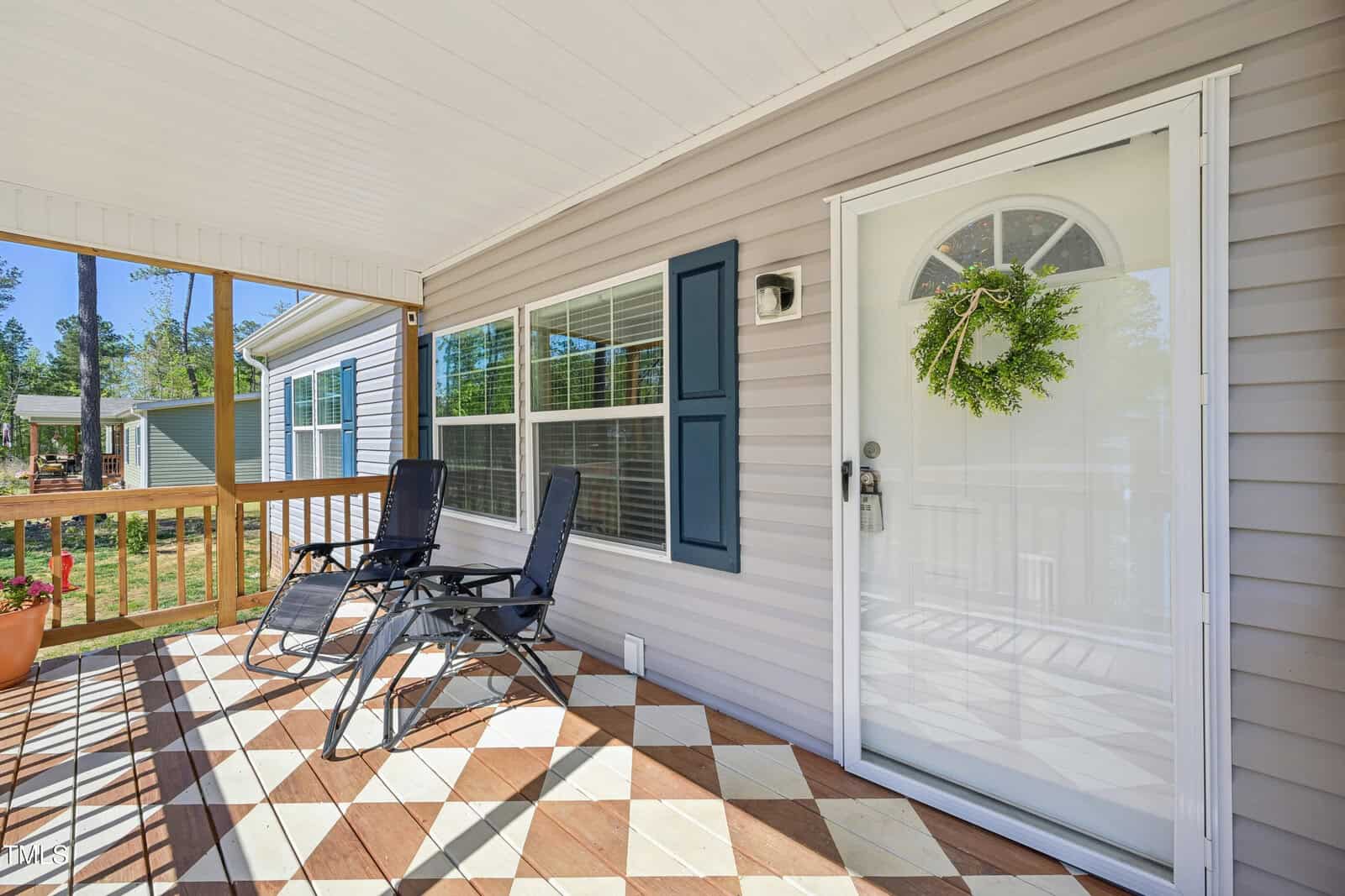 A covered porch with two black lounge chairs, a potted plant, and a glass door decorated with a green wreath. The porch floor has a diamond pattern, and the house has beige siding with blue shutters.