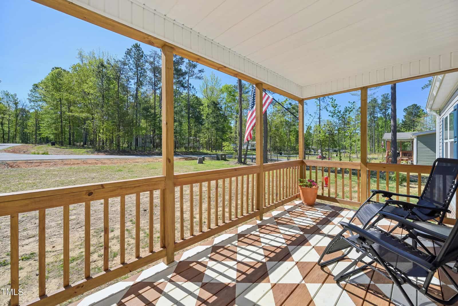 A covered wooden porch with railing, two black chairs, a potted plant, and an American flag. The porch overlooks a yard with trees and a dirt road under a clear blue sky.