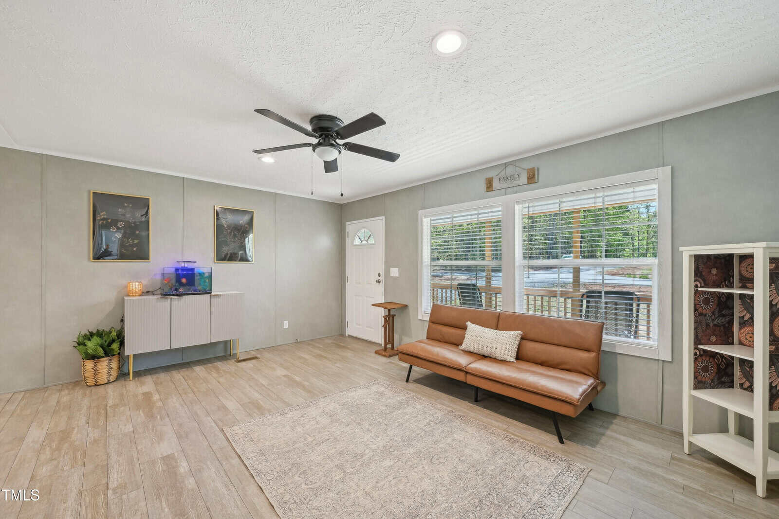 A living room with light wood flooring, a brown leather sofa, a neutral area rug, a ceiling fan, wall art, a small cabinet with plants and an aquarium, and large windows with blinds letting in natural light.