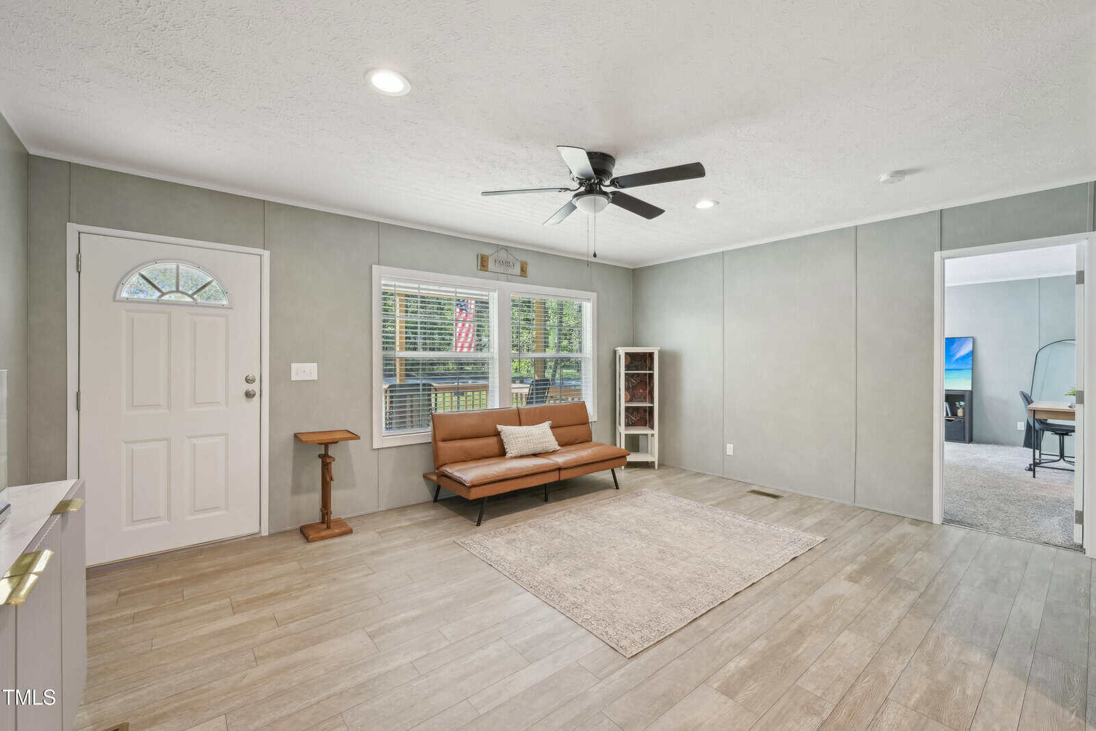 A bright living room with a ceiling fan, wood-look tile floor, a brown futon, a small rug, a cat tree, a bookshelf, and a window showing an American flag outside. Adjoining room has a TV and desk.