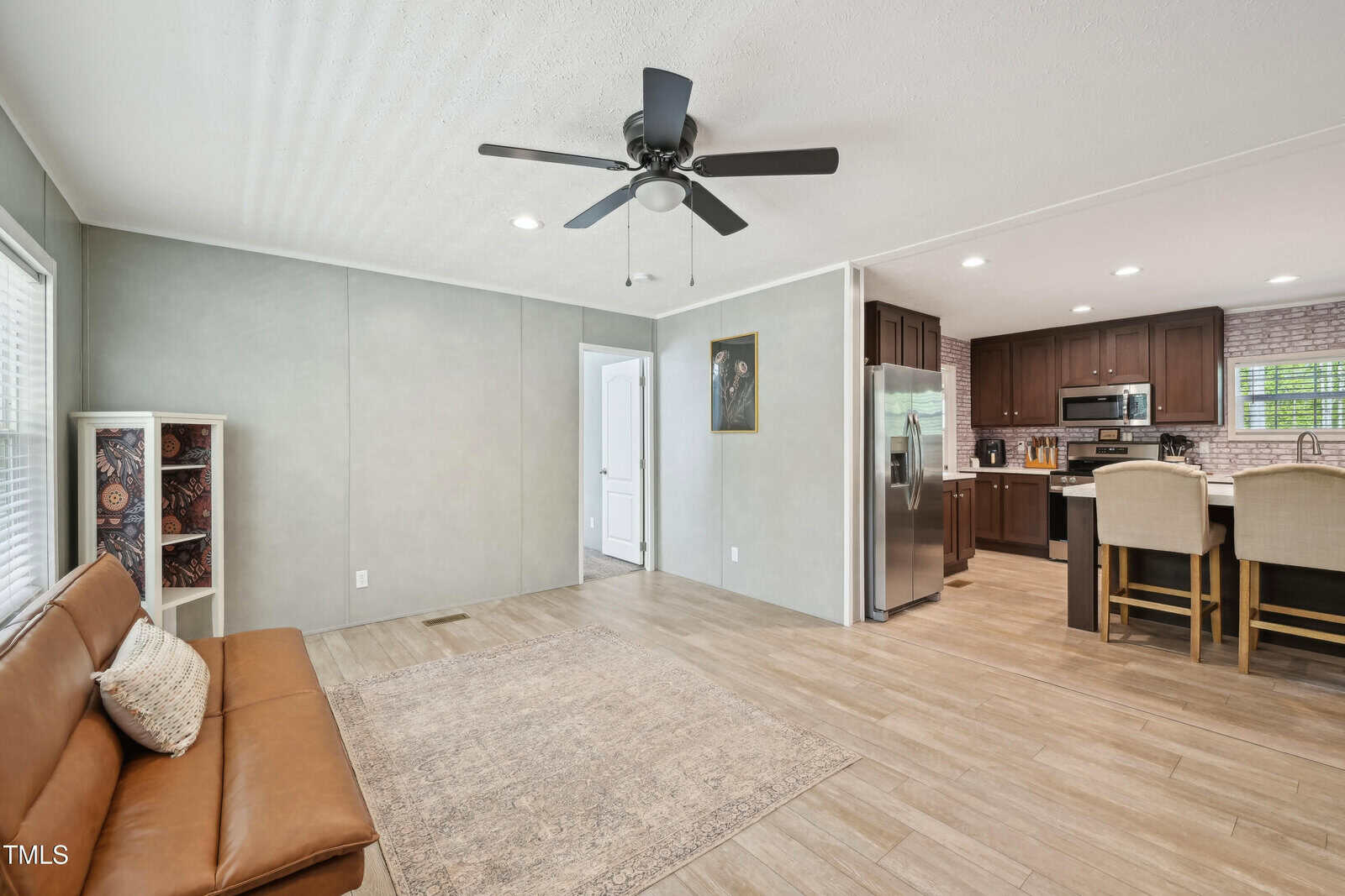 A living area with a tan leather sofa, light area rug, and ceiling fan; adjacent to an open kitchen with dark wood cabinets, stainless steel appliances, and a breakfast bar with upholstered chairs.
