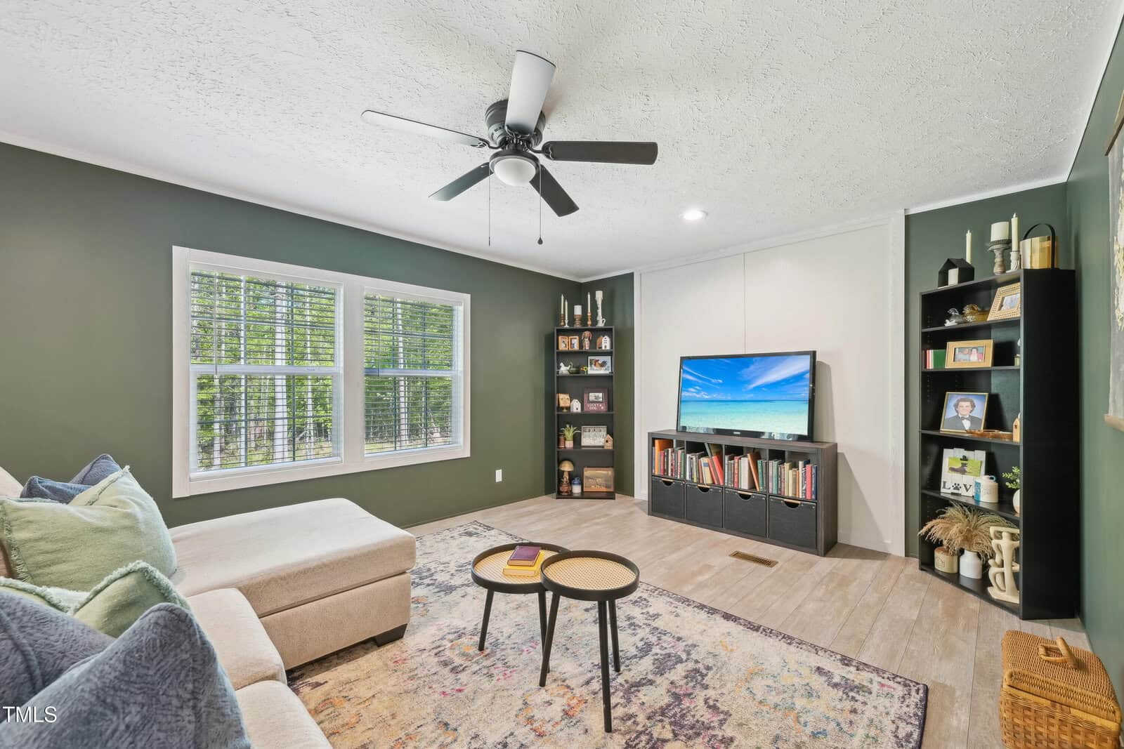 A living room with a light-colored sectional sofa, two round coffee tables, a wall-mounted TV, two black bookshelves, green walls, a ceiling fan, and a window overlooking trees.