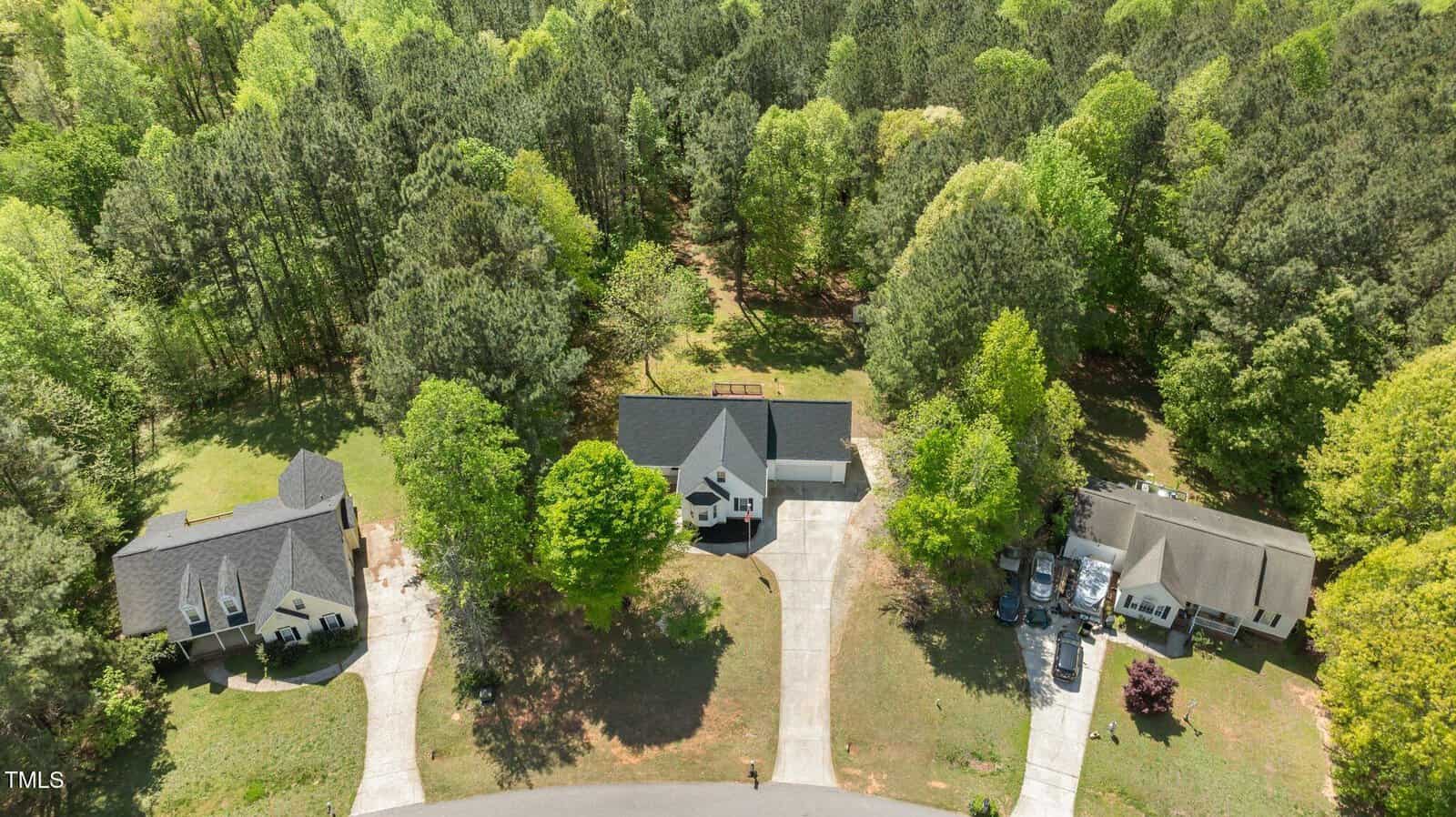 Aerial view of three suburban homes with driveways, surrounded by tall pine trees and greenery at the end of a cul-de-sac, bordering a wooded area in the background.