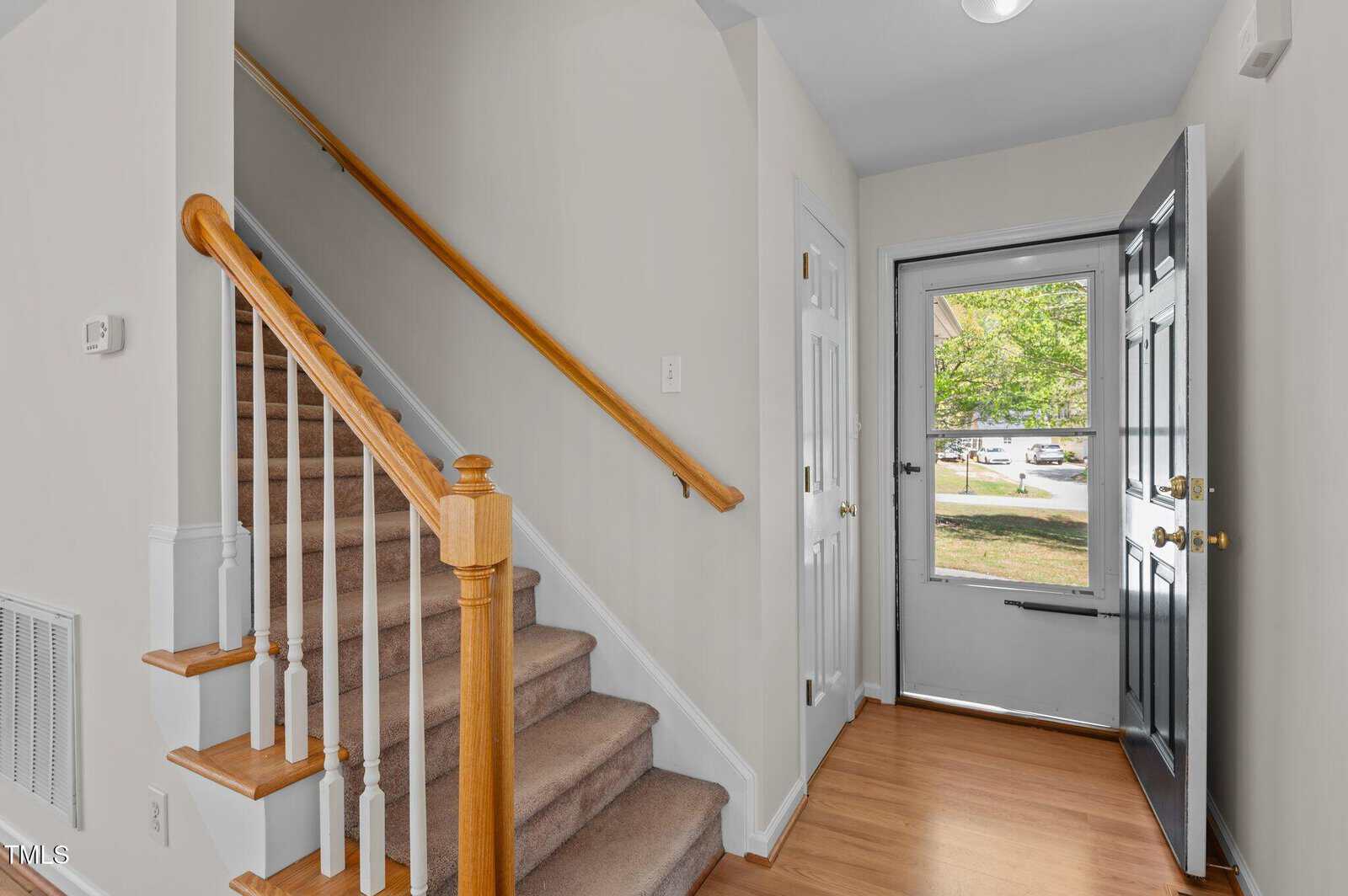 A view of a home's entryway with an open black front door, a wooden staircase with white spindles and beige carpet, hardwood flooring, and white walls. Sunlight is visible outside through the storm door.