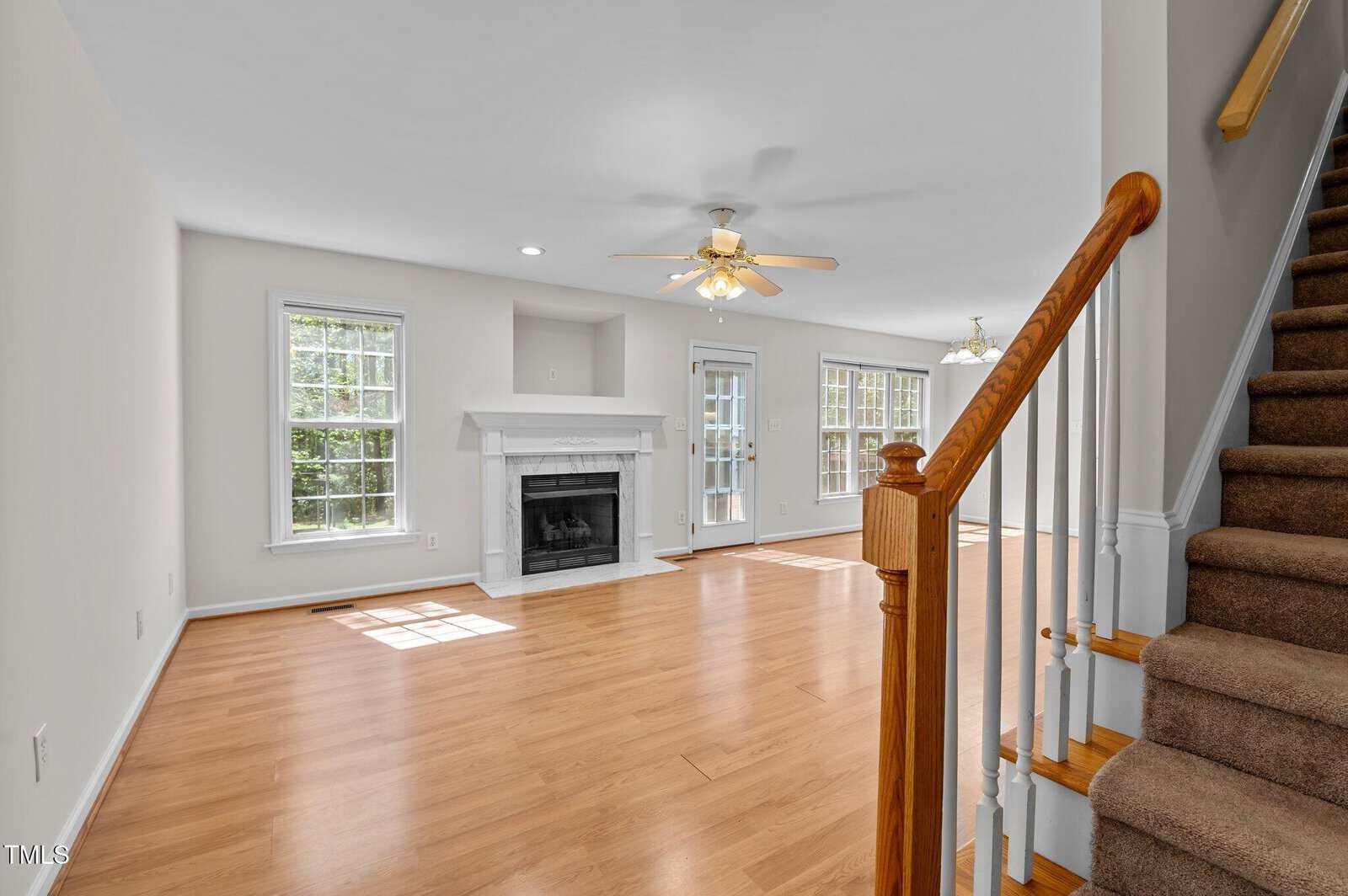 A bright living room with light wood floors, white walls, a fireplace, ceiling fan, two large windows, glass door, and a carpeted staircase with a wooden railing leading to the upper floor.