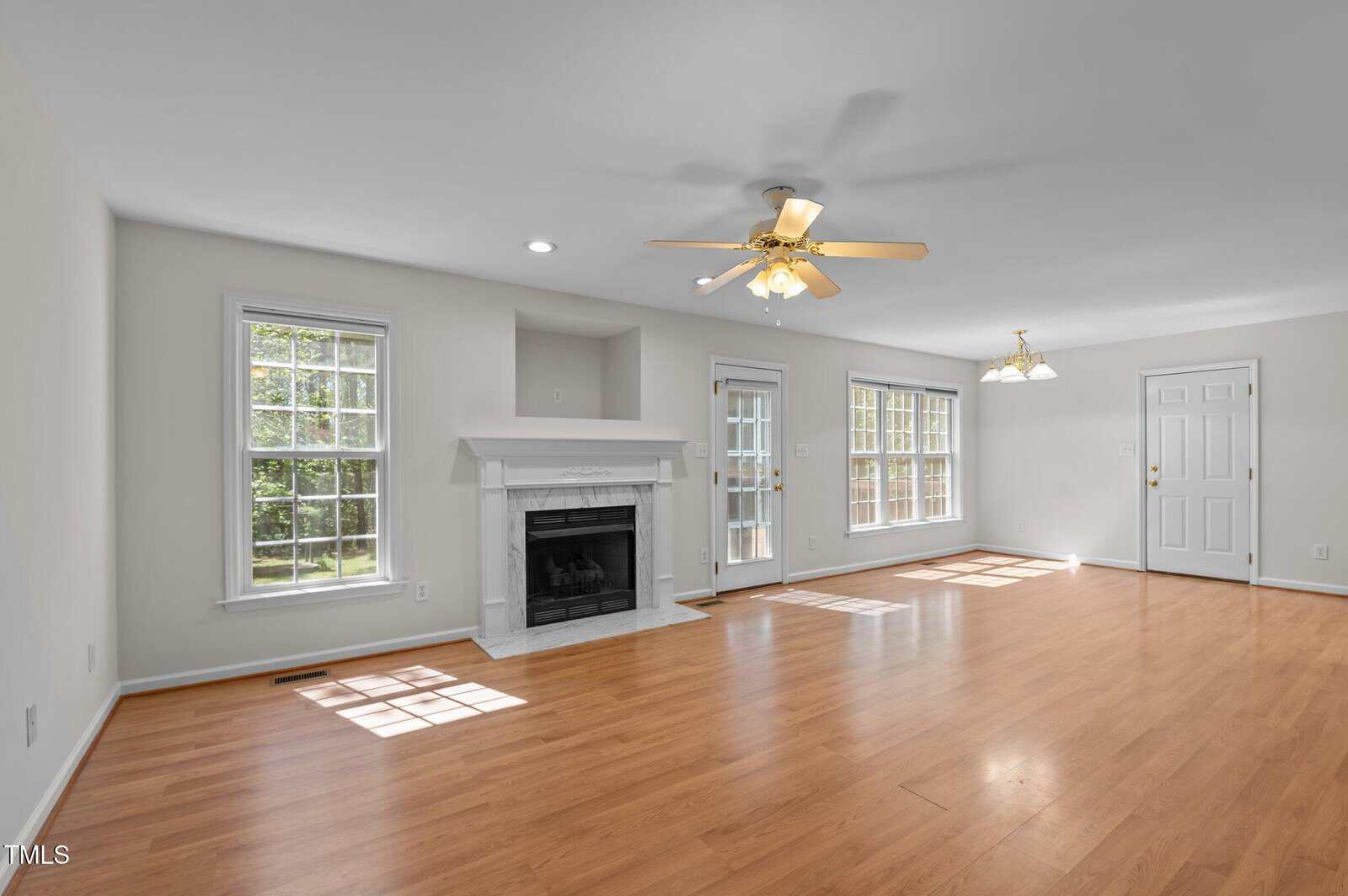 Spacious living room with light wood floors, white walls, a ceiling fan, a fireplace with a white mantel, large windows, glass door to outside, and a white entry door. Room is brightly lit by natural light.
