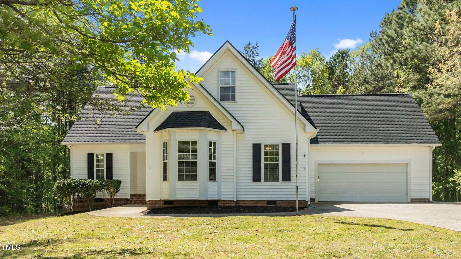 A white, single-story house with black shutters and a two-car garage is shown. An American flag is mounted near the front. Trees and greenery surround the house, and the sky is clear.