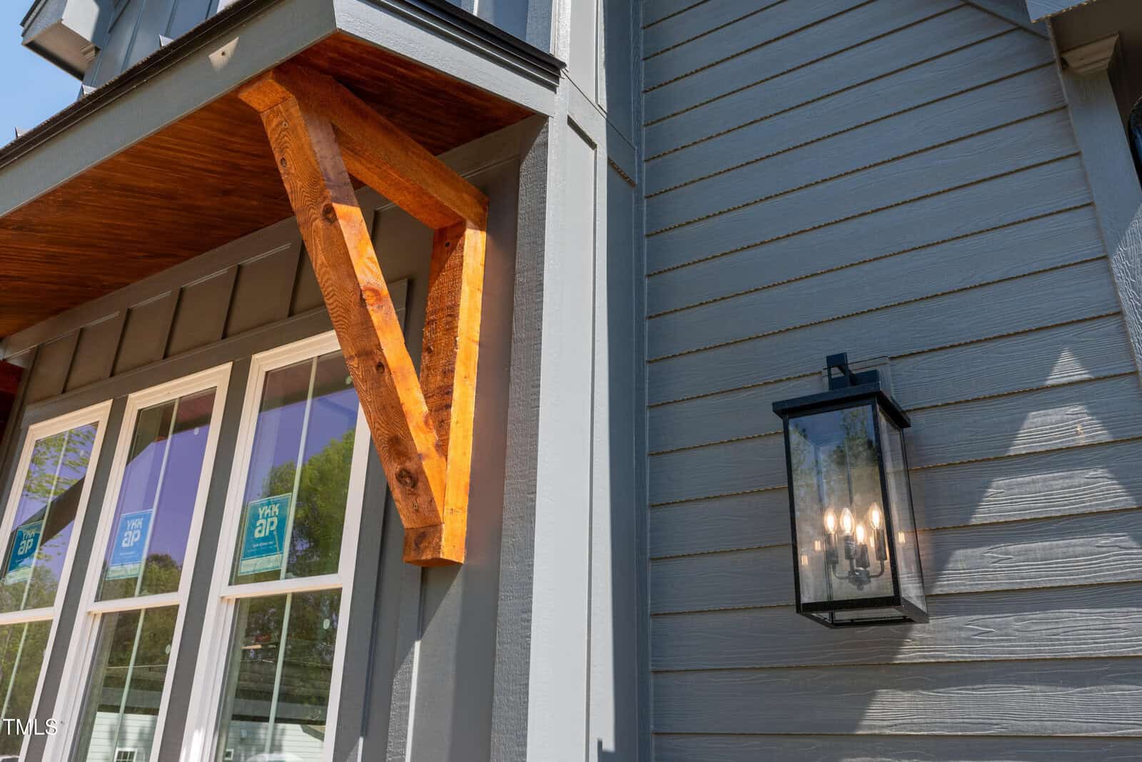 Close-up of a house exterior showing gray vertical siding, a wooden decorative bracket above windows with stickers, and a black wall-mounted lantern-style light fixture.