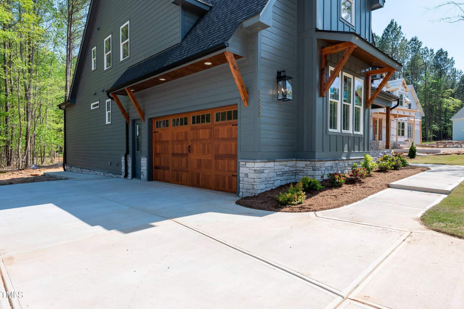 A modern house with dark gray siding, a wooden garage door, stone accents, and a large concrete driveway surrounded by landscaped mulch and plants. Another house is visible in the background.