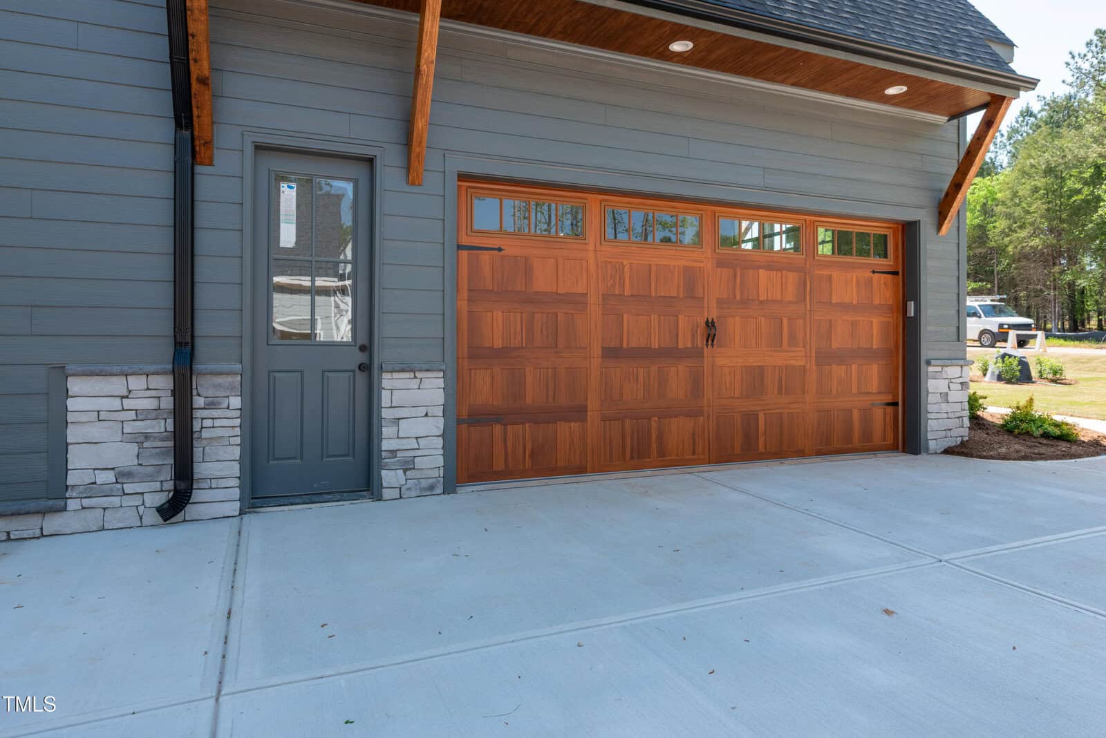 A modern two-car garage with a wooden overhead door, a gray side door, stone accents on the lower wall, and a smooth concrete driveway. Green trees and a white vehicle are visible in the background.