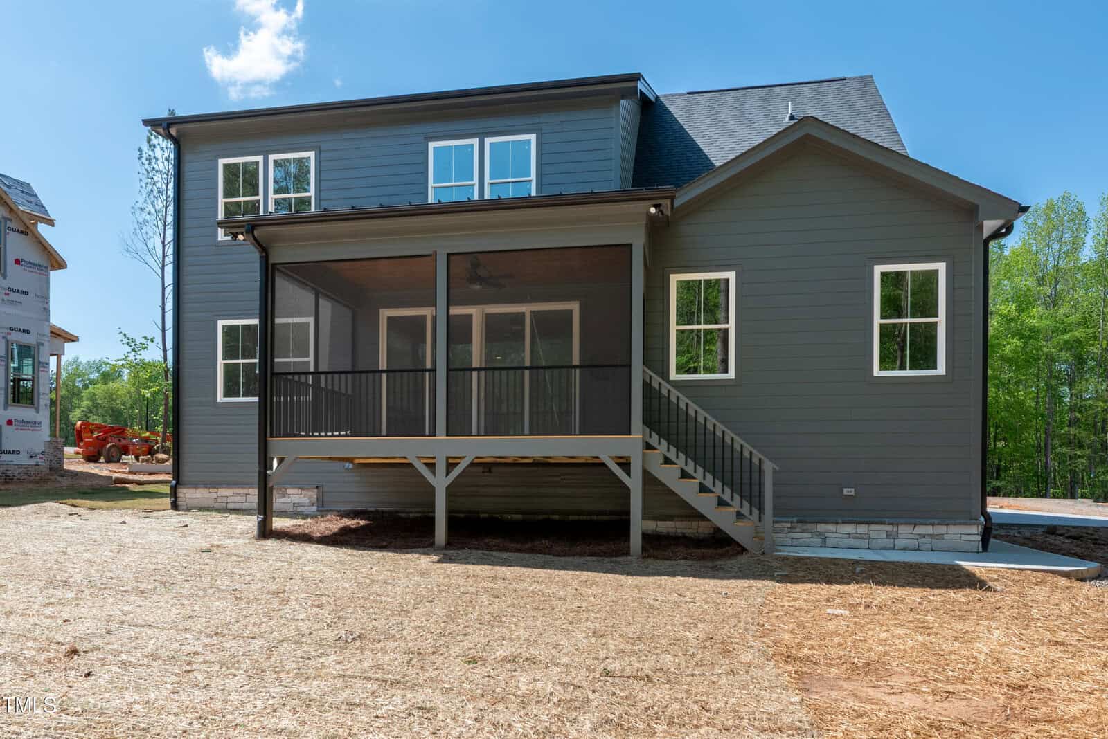 A newly constructed two-story house with gray siding, white trim, and a screened-in back porch elevated on wooden posts. The yard is covered with straw, and trees are visible in the background.