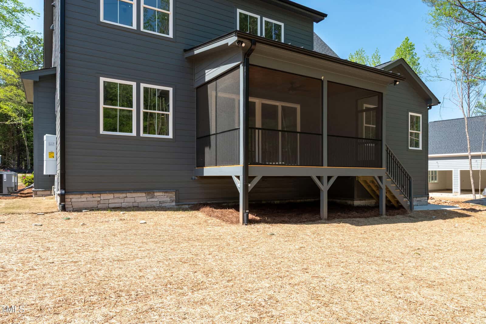 A screened-in porch with steps leads up to the back entrance of a two-story dark gray house with white trim. The yard is covered with straw and has no grass. Another house is visible in the background.