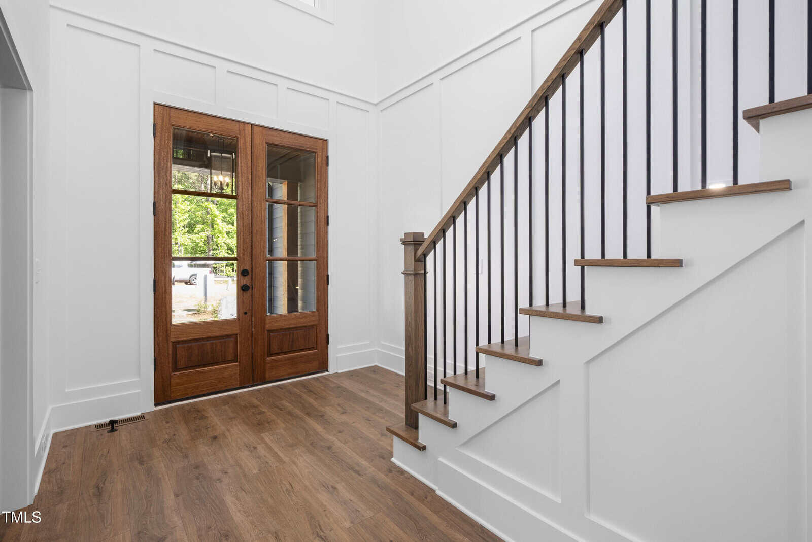 A modern entryway with light wood flooring, white paneled walls, a wooden staircase with black metal railings, and double glass front doors with wood frames. Natural light comes in through the doors.