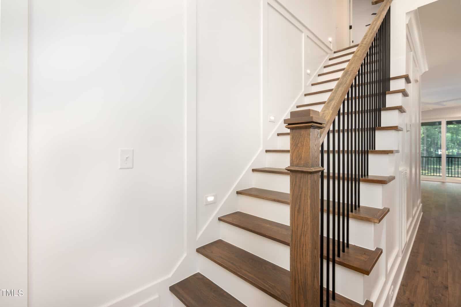 A wooden staircase with dark handrails and black balusters leads to the upper floor of a modern home with white walls and wood flooring. Natural light comes from the windows in the background.