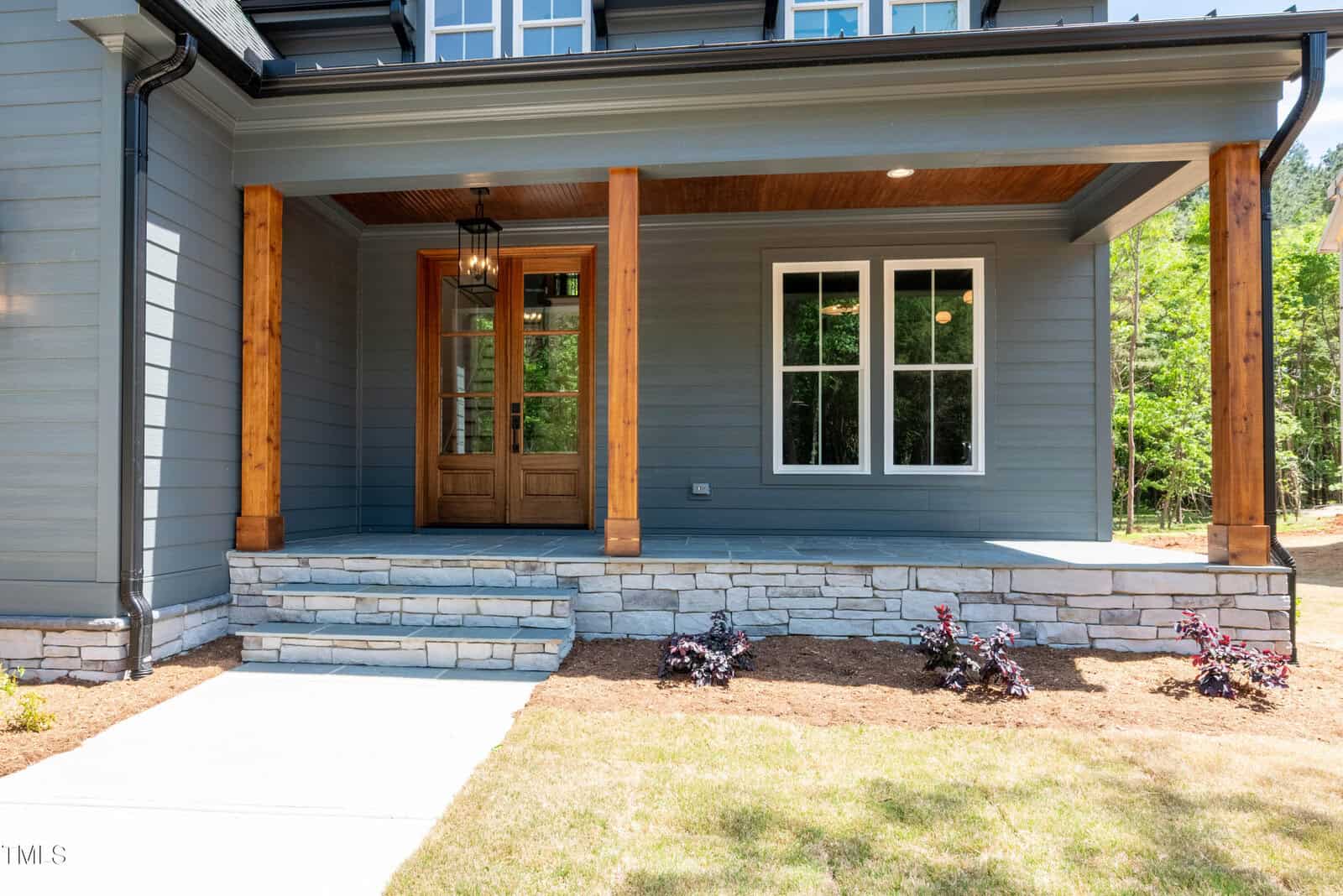 A modern house front porch with wooden columns, stone steps, gray siding, a wooden front door with glass panels, and two white-framed windows. Small plants are planted along the border of the porch.