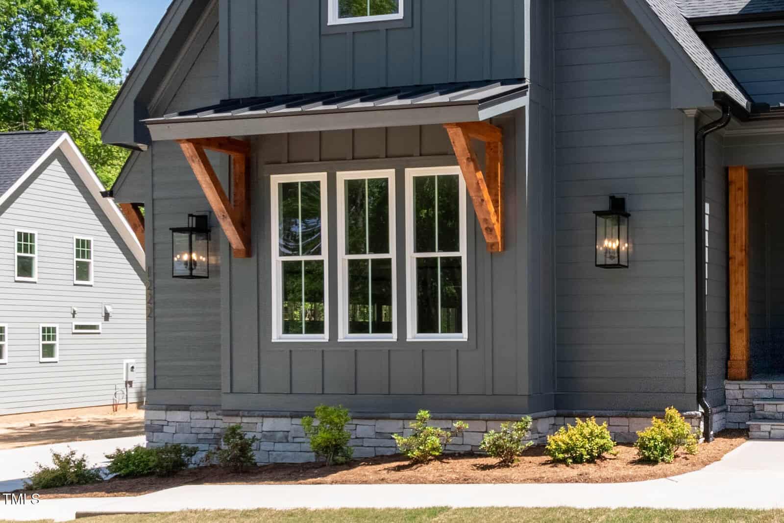 A close-up of a modern house exterior with gray vertical siding, a large white-framed window, wooden awnings, two black wall lanterns, stonework along the foundation, and landscaped shrubs in front.