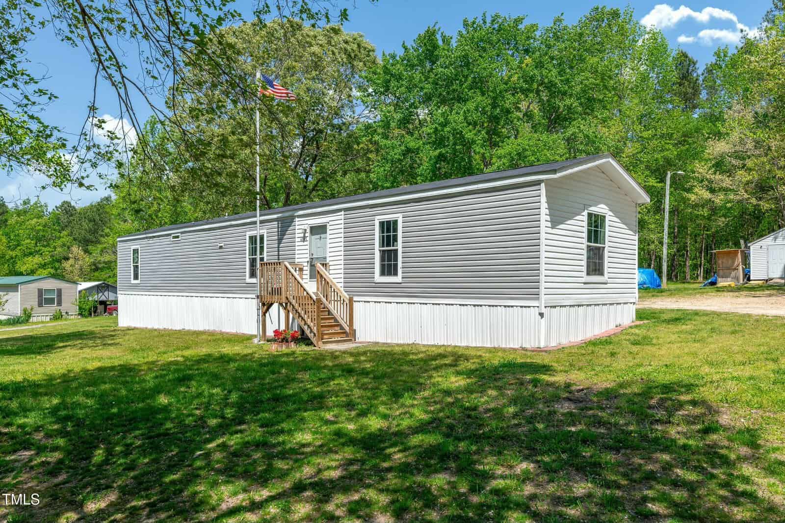A single-wide manufactured home with gray siding and white trim sits on a grassy lot. A small wooden porch with stairs leads to the front door. Trees and an American flag are visible in the background.