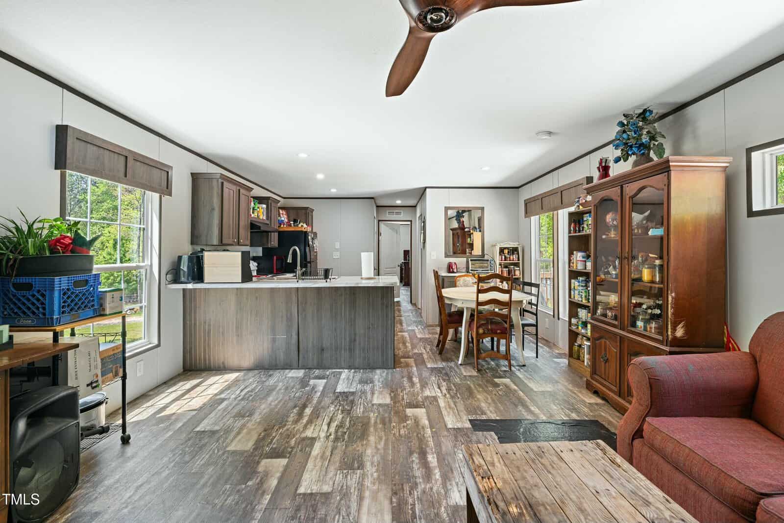 A spacious open-concept kitchen and dining area features wood-style flooring, a central island with stools, a dining table, a china cabinet with glass doors, and a red armchair in the foreground.