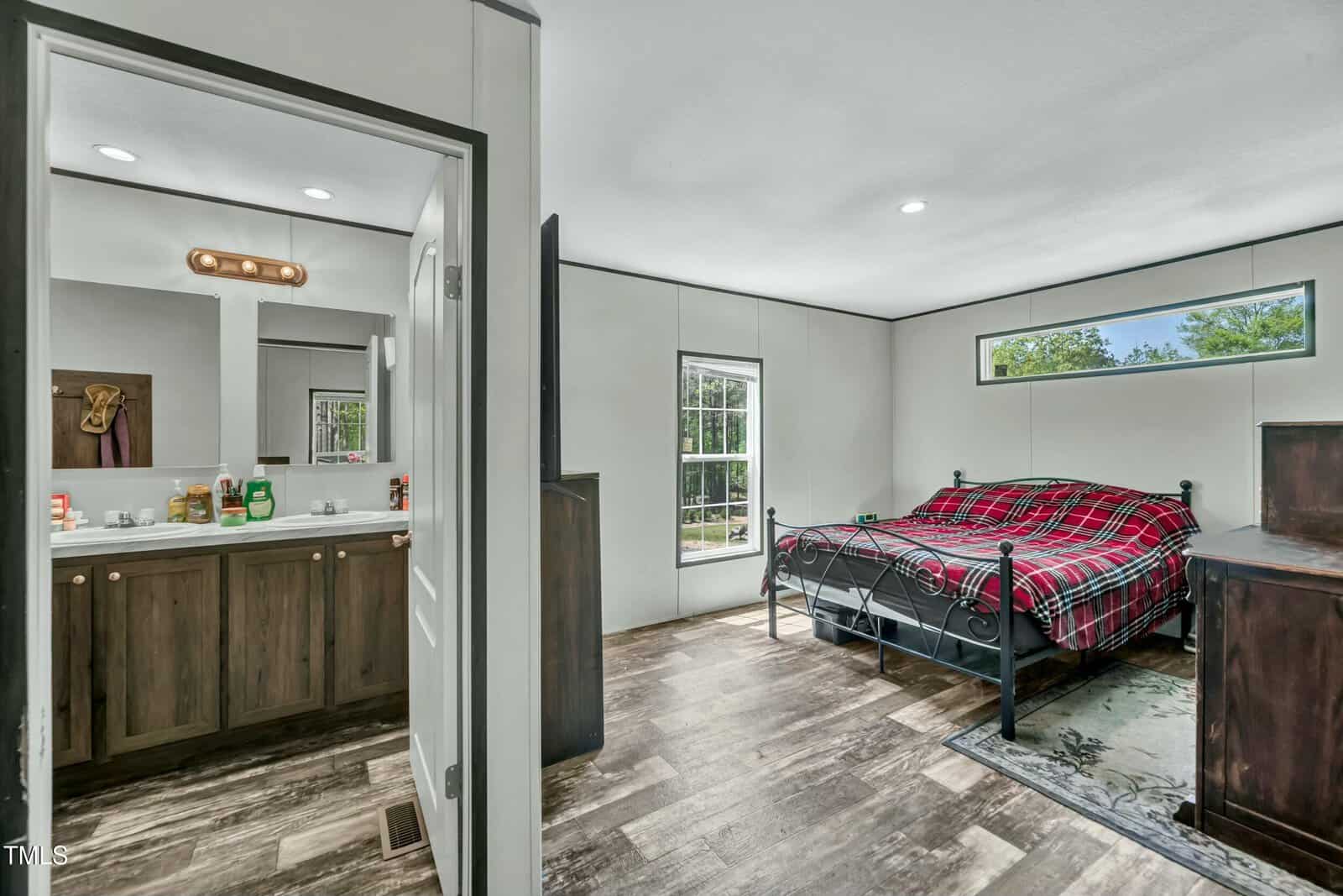 A bedroom with a metal-framed bed covered in a red plaid blanket, a wooden dresser, and a window; an adjacent bathroom with a double sink vanity and mirror is visible through an open door.