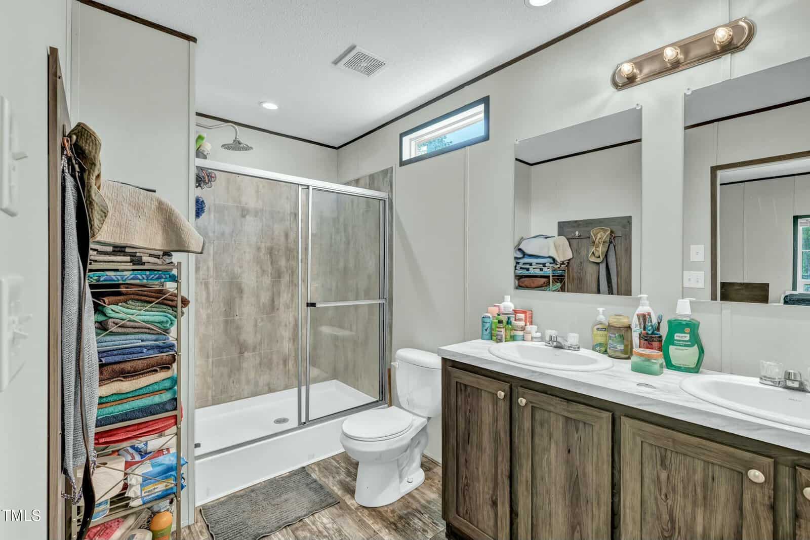 A bathroom with a glass-enclosed shower, toilet, double vanity with two mirrors, and various toiletries on the counter. A tall shelving unit on the left holds neatly folded towels and other items.