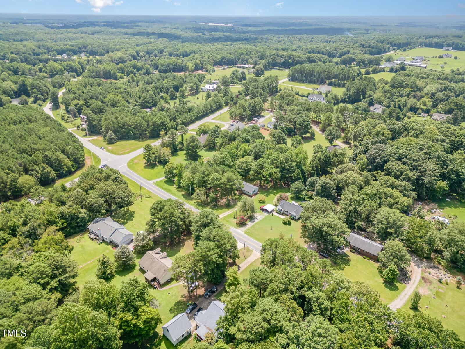 Aerial view of a rural neighborhood with houses, roads, and abundant green trees and lawns, surrounded by dense forest and open fields under a partly cloudy sky.