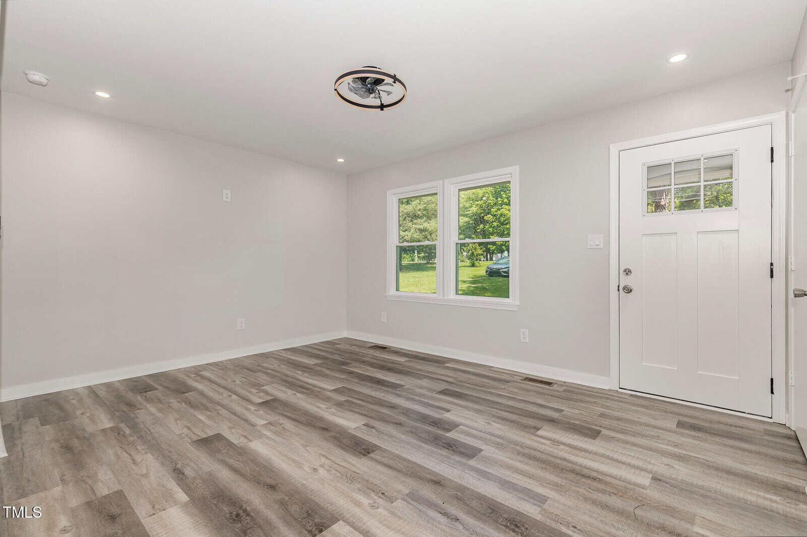 Empty room with light gray walls, wood-look vinyl flooring, two windows, a white door with glass panels, ceiling vent, and recessed lighting. Tree-filled greenery is visible outside through the windows.