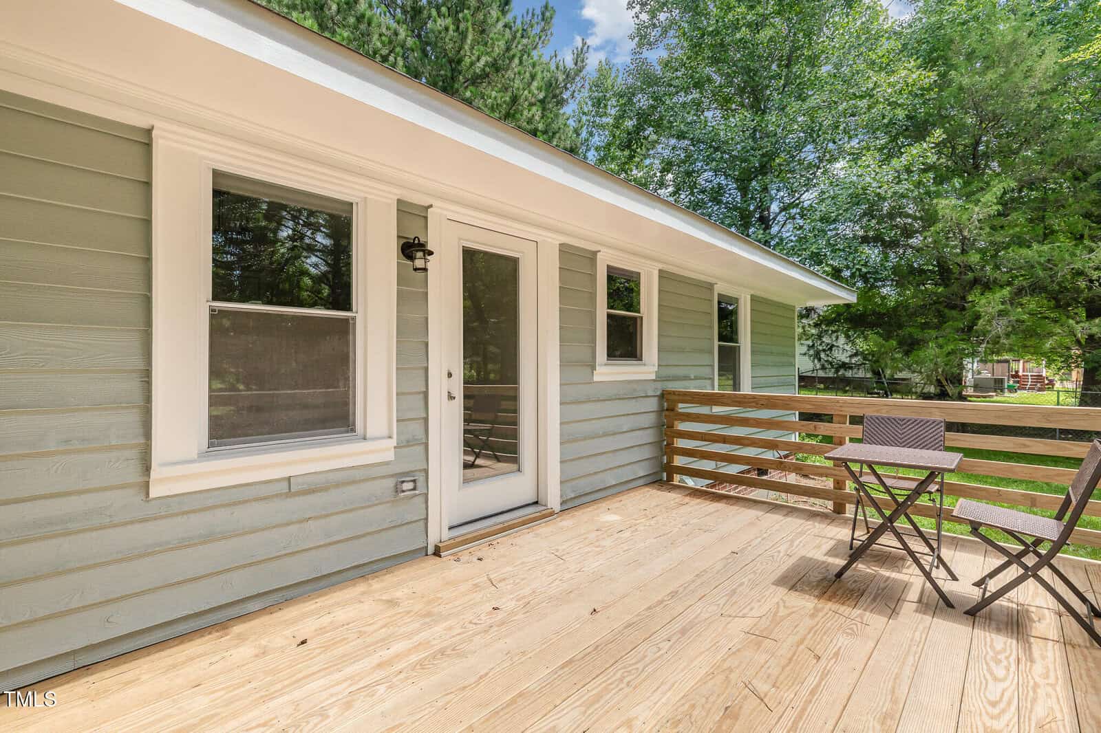 A wooden deck with a small table and two chairs, attached to a house with light blue siding and white trim. There are trees and greenery visible in the background.